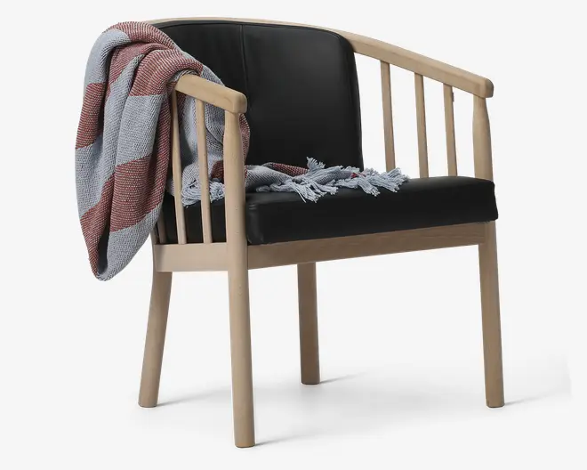 Stol med lyst træstel og sort polstret sæde og ryglæn. Stolen har lodrette tremmer på ryg og sider, og et rød- og hvidstribet tæppe med frynser er lagt over det ene armlæn.