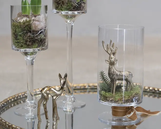 Klare dekorative glasgenstande arrangeret på et spejlfad, hver med et lille skovmotiv med mos, små sten og metalliske hjortefigurer i høje glas på stilk og en cylindrisk glasbeholder.