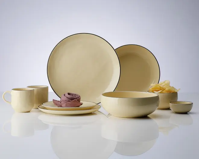 Et sæt cremefarvet keramikservice med blank overflade, bestående af to store tallerkener, en mellemstor skål, en lille skål, to krus og en sidetallerken. Delene har en diskret mørk kant og et glat, minimalistisk design. Sættet er arrangeret på en reflekterende overflade, hvilket fremhæver dets elegante og moderne udseende.