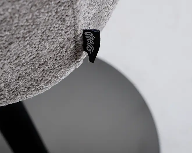 Nærbillede af en tekstureret, lysegrå stofoverflade med et lille sort stofmærke fastgjort, der har hvid, stiliseret tekst. Stoffet ser blødt og plysset ud, og syningen er synlig langs sømmen. Baggrunden er uskarp og viser en glat, rund overflade i en neutral farve.