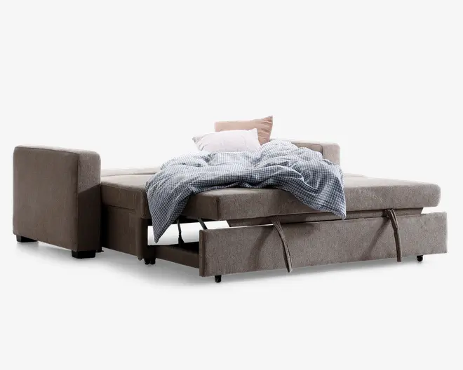 Sovesofa i en neutral beige farve, vist i udfoldet sengeposition. Designet har en udtrækssektion med stofhåndtag, der gør det nemt at omdanne sofaen til en seng. Sengen er dækket med et lyseblåt ternet tæppe og to puder, én hvid og én lyserød. Betrækket ser blødt ud, og stilen er moderne og funktionel, velegnet til små rum eller gæsteværelser.