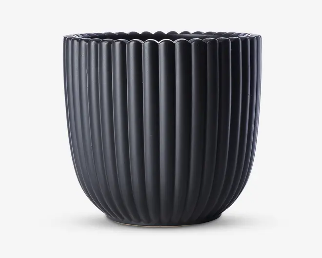 En mat sort urtepotteskjuler med lodrette riller og en rund, let tilspidset form. Kanten har en blød bølget kant, som giver et dekorativt udtryk. Overfladen er glat, og potten ser ud til at være lavet af keramik.