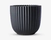 En mat sort urtepotteskjuler med lodrette riller og en rund, let tilspidset form. Kanten har en blød bølget kant, som giver et dekorativt udtryk. Overfladen er glat, og potten ser ud til at være lavet af keramik.