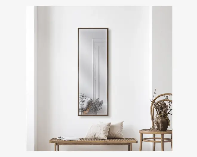 Rektangulært vægspejl med en smal, mørk ramme, monteret lodret på en hvid væg. Spejlet reflekterer et minimalistisk interiør med en dør og nogle dekorative tørrede planter i vaser. Opsætningen inkluderer en træbænk med to lyse puder og en bog samt en rund træstol med en vase med tørrede blomster, hvilket skaber en naturlig og hyggelig stemning.