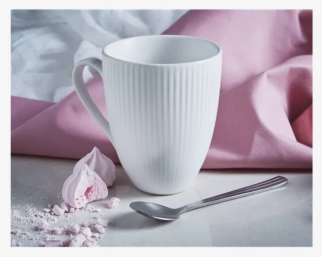 Høj, hvid krus med lodret rillet tekstur, stort buet hank og glat, blank overflade. Kruset står ved siden af en metalske og nogle lyserøde marengsstykker på en lys overflade, med et blødt lyserødt stof i baggrunden.