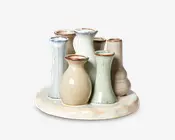 Et sæt med syv små keramikvaser, hver med sin egen unikke form og forskellige højder, arrangeret sammen på en rund, glaseret keramikbase. Vaserne har bløde, jordnære nuancer som beige, lyseblå, lysegrøn og offwhite, med en blank finish og diskret plettet tekstur. Vaskanten er en smule mørkere, hvilket giver et håndlavet, kunsthåndværkspræg.