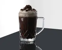 Et klart glas med solidt håndtag, fyldt med en mørk, chokoladeagtig drik. Drikken er toppet med et generøst lag flødeskum og drysset med chokoladekiks-stykker. Glasset har et dekorativt, facetslebet mønster i bunden, som giver et elegant udtryk.