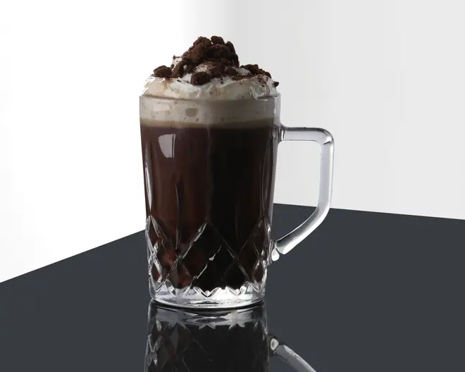 Et klart glas med solidt håndtag, fyldt med en mørk, chokoladeagtig drik. Drikken er toppet med et generøst lag flødeskum og drysset med chokoladekiks-stykker. Glasset har et dekorativt, facetslebet mønster i bunden, som giver et elegant udtryk.