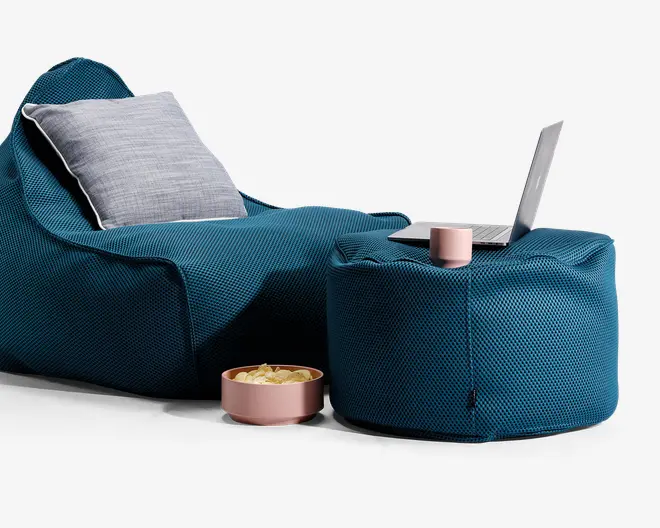 Billedet viser et moderne siddearrangement med en dybblå, tekstureret sækkestol og en lysegrå, rektangulær pude for ekstra komfort. Ved siden af står en matchende blå puf, også med tekstur, som fungerer som et sidebord. På puffen står en lyserød kop og en åben bærbar computer, hvilket indikerer et arbejds- eller afslapningsområde. Foran stolen er der en lyserød skål med kartoffelchips, hvilket giver et afslappet og hyggeligt miljø. Materialerne ser bløde og slidstærke ud, velegnet til afslapning eller arbejde.