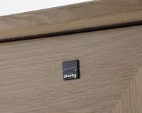 Nærbillede af en lys egetræsoverflade med et lille, kvadratisk, sort metalmærke med mærkenavnet 'Skovby' i hvide bogstaver. Træet har en naturlig årestruktur og mat finish, og der ses en skrå kantdetalje øverst.