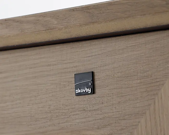 Nærbillede af en lys egetræsoverflade med et lille, kvadratisk, sort metalmærke med mærkenavnet 'Skovby' i hvide bogstaver. Træet har en naturlig årestruktur og mat finish, og der ses en skrå kantdetalje øverst.