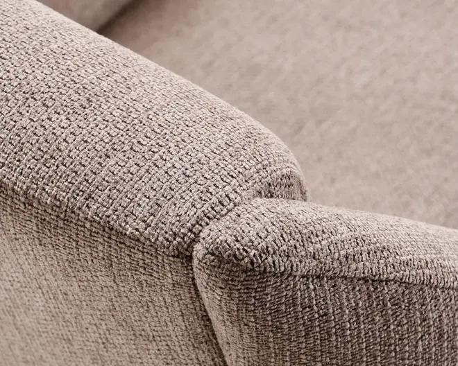 Nærbillede af en sofa-armlæn med tekstureret, vævet stof i en neutral beige farve. Polstringen har et diskret ternet mønster, som giver en blød og slidstærk overflade. Armlænet er let afrundet i kanterne, hvilket giver et moderne og komfortabelt udtryk.