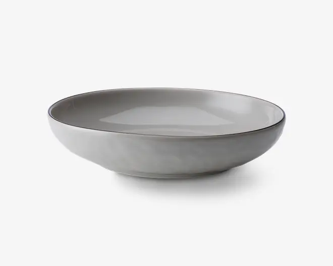 En rund, lav skål med en glat, mat lysegrå yderside og en blank inderside. Kanten er let forhøjet og har en diskret mørkere nuance, hvilket giver en elegant kontrast. Skålen har et minimalistisk, moderne design med rene linjer og en blødt buet form.