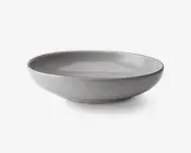 En rund, lav skål med en glat, mat lysegrå yderside og en blank inderside. Kanten er let forhøjet og har en diskret mørkere nuance, hvilket giver en elegant kontrast. Skålen har et minimalistisk, moderne design med rene linjer og en blødt buet form.