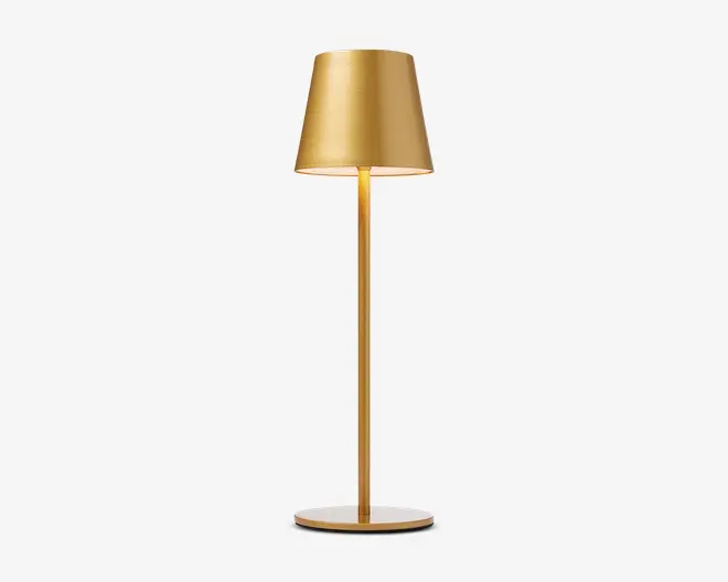 Elegant, moderne lampe med mat guld-finish. Lampen har en konisk lampeskærm, en slank cylindrisk stang og en flad, rund fod. Designet er minimalistisk og stilfuldt, velegnet til moderne indretning.