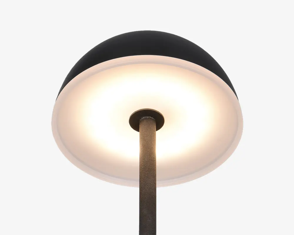 Nærbillede af en lampe med en bred, kuppelformet skærm. Lampens yderside er mat sort, mens indersiden er hvid og reflekterer et varmt, diffust lys. Lampestangen er metal med en mørk, struktureret overflade, og designet er moderne og minimalistisk.