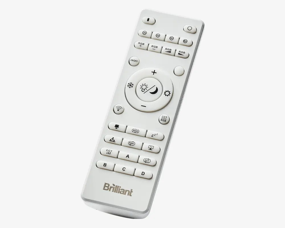 Hvid fjernbetjening med mange knapper, herunder dedikerede knapper til RGB-farveindstillinger, justering af lysstyrke og lysindstillinger som dagslys og nattilstand. Har mærkede knapper til gruppekontrol (A, B, C, D), timerfunktioner og en stor central cirkulær knap til lysstyrke og tilstand. 'Brilliant'-logoet er trykt nederst.