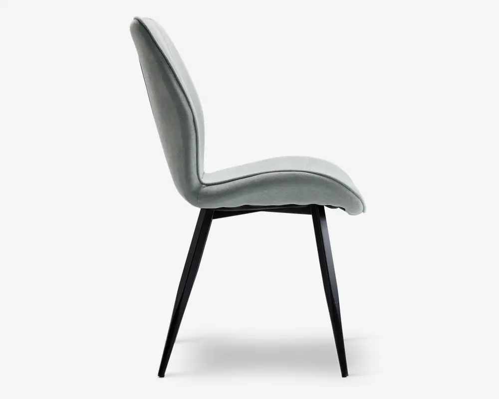 Sidevisning af en moderne stol med let buet, polstret ryglæn og sæde betrukket med et blødt, lysegråt stof. Stolen har slanke, skråtstillede sorte metalben, der giver et stilrent og minimalistisk udtryk. Sæde og ryglæn er sømløst forbundet og giver ergonomisk støtte samt et moderne design.