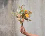 En hånd holder en lille buket med en blanding af tørrede blomster og løv. Buketten indeholder gule og ferskenfarvede blomster, fine kviste og blade med forskellige strukturer. Blomsterne har et rustikt og naturligt udseende, og stilkene er samlet uden synlig indpakning.