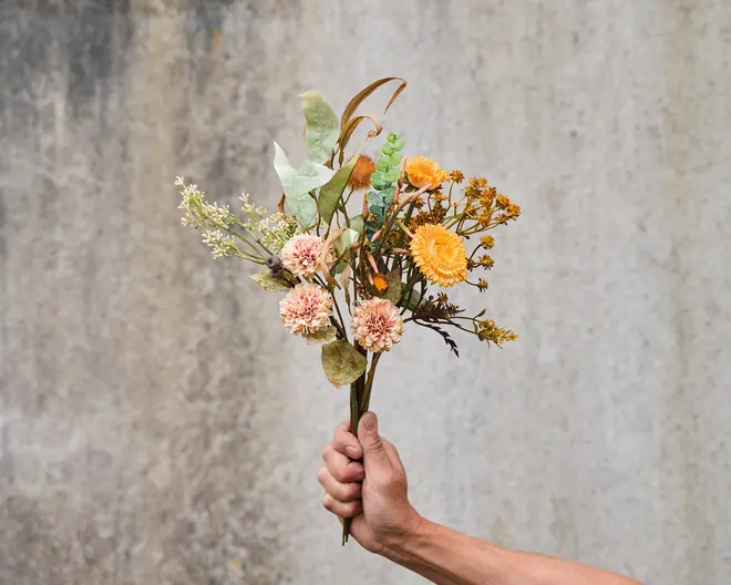 En hånd holder en lille buket med en blanding af tørrede blomster og løv. Buketten indeholder gule og ferskenfarvede blomster, fine kviste og blade med forskellige strukturer. Blomsterne har et rustikt og naturligt udseende, og stilkene er samlet uden synlig indpakning.