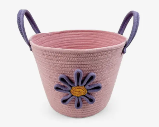 En rund, lyserød flettet kurv med to solide lilla håndtag. Kurven har en dekorativ blomst på forsiden, lavet af lilla kronblade og en gul midte, hvilket giver et legende og muntert udtryk. Kurven har en glat, snoet tekstur og en bred åbning for nem adgang.