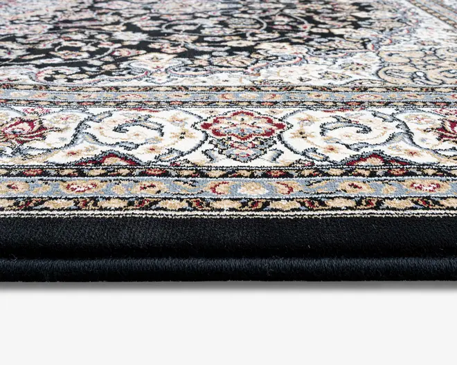 Nærbillede af et tykt, blødt tæppe med et detaljeret, ornamenteret mønster. Tæppet har en sort baggrund med indviklede blomster- og snirklede motiver i creme, beige, rød og lyseblå. Kanten er dekoreret med symmetriske designs og flere accentfarver, hvilket giver et luksuriøst og elegant udseende. Luven ser tæt og blød ud, hvilket indikerer komfort og holdbarhed.