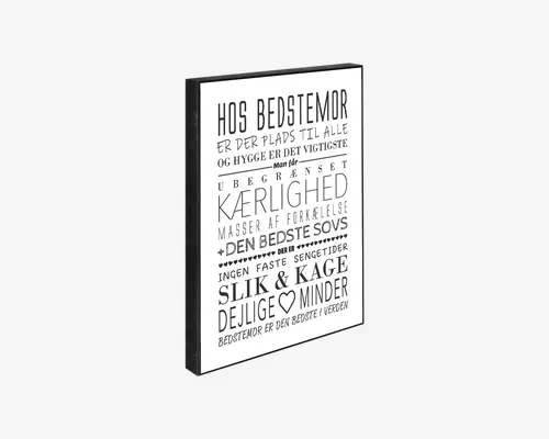 Rektangulært vægkunst med sort ramme og hvid baggrund, der viser forskellige danske tekster i varierende skrifttyper og størrelser. Teksten hylder en bedstemor og nævner ubegrænset kærlighed, den bedste sovs, slik og kage, hygge og dejlige minder. Designet bruger legende typografi, både fede og almindelige skrifttyper, og har et lille hjerte som dekoration.