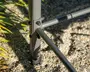 Nærbillede af et robust metalstel-samling med tre rørformede stænger, der er forbundet med bolte og møtrikker. Stellet er malet gråt og har en sort plastikfod nederst for stabilitet. Samlingspunkterne er forstærket med ekstra beslag, hvilket indikerer holdbarhed og sikker montering. Stellet står på en grusbelagt overflade udendørs, med noget grønt løv synligt i baggrunden.