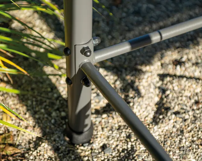Nærbillede af et robust metalstel-samling med tre rørformede stænger, der er forbundet med bolte og møtrikker. Stellet er malet gråt og har en sort plastikfod nederst for stabilitet. Samlingspunkterne er forstærket med ekstra beslag, hvilket indikerer holdbarhed og sikker montering. Stellet står på en grusbelagt overflade udendørs, med noget grønt løv synligt i baggrunden.