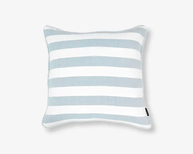 Firkantet pude med brede, horisontale striber i skiftevis lyseblå og hvide farver. Kanterne er pænt afsluttet med en diskret piping, og der sidder et lille sort stofmærke på den ene side. Stoffet ser vævet ud, hvilket giver puden et blødt og tekstureret udtryk.