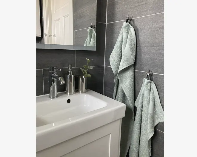 To lysegrønne håndklæder med en diskret tekstureret kant hænger på kromkroge mod en mørkegrå flisevæg på badeværelset. Håndklæderne ser bløde og fyldige ud, hvor det ene er større end det andet. Billedet viser også en hvid vask med kromarmatur, en sæbedispenser og en lille plante i en metalbeholder, hvilket giver et moderne og rent udtryk.