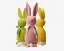 Tre høje, stiliserede kaninfigurer i pastelfarver – lyserød, grøn og gul – vises. Hver kanin har en glat, mat overflade og lange ører. Den lyserøde kanin foran har et hvidt bånd bundet i en sløjfe om halsen, den grønne har et grønt bånd, og den gule har ikke noget synligt bånd. Der ligger stykker af knækkede hvide chokoladeæg ved deres fødder, hvilket antyder, at de er lavet af chokolade.