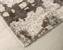 Nærbillede af et tykt, blødt tæppe med et moderne abstrakt mønster. Tæppet har en blanding af grå, creme og diskrete rust-orange nuancer, hvilket giver et tekstureret, plettet udseende. Kanterne er pænt afsluttede med en tæt kant, og overfladen ser blød og tæt ud, hvilket giver god komfort under fødderne.