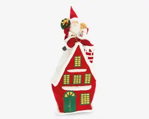 En festlig dekoration med et blødt rødt hus, der har en grøn dør og vinduer med gule ruder, alt sammen kantet med hvidt for at ligne sne. På det snedækkede tag sidder en blød julemandsfigur med en sæk gaver og en bamse, benene hænger ud over kanten, og der er en skorsten ved siden af ham. Udtrykket er farverigt og hyggeligt, ideelt til juleudsmykning.