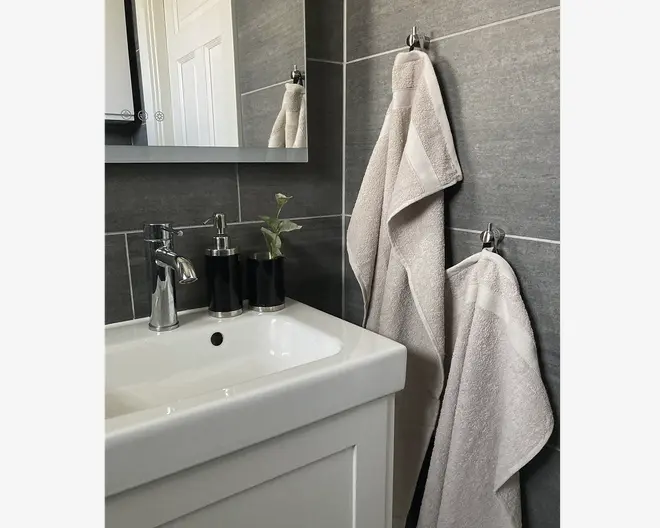 To lysegrå håndklæder med en diskret kantdetalje hænger på kromkroge mod en mørkegrå flisevæg på badeværelset. Håndklæderne ser bløde og fyldige ud med pænt afsluttede kanter. Det større håndklæde hænger øverst, mens det mindre hænger nedenunder. Håndklæderne passer til det moderne, minimalistiske badeværelsesdesign.