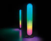 To vertikale, ovale LED-lysstave med afrundede toppe står på cirkulære baser. Lysene viser en glidende overgang af klare farver, herunder blå, grøn, gul og pink, som smelter sammen i et blødt farveskift. Overfladen under lysene reflekterer det farverige skær, og baggrunden er helt sort, hvilket fremhæver lysenes klare og farverige effekter.