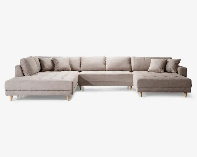 Stor L-formet sofa med lyse beige stofbetræk, polstrede sæder med knapdetaljer og lav ryg. Sofaen har flere ryg- og sidepuder for ekstra komfort og står på korte, skrå træben. Designet er moderne og rummeligt, velegnet til at rumme flere personer.