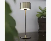 En moderne bordlampe med børstet metalfinish, cylindrisk lampeskærm og en slank, lige stang, der forbinder til en rund, flad fod. Lampen udsender et blødt, varmt lys under skærmen og skaber en hyggelig stemning. Det minimalistiske design passer godt til moderne indretning.