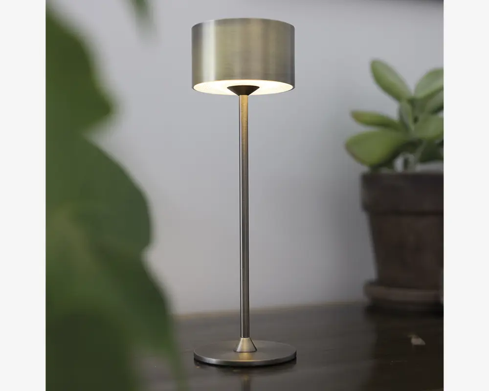 En moderne bordlampe med børstet metalfinish, cylindrisk lampeskærm og en slank, lige stang, der forbinder til en rund, flad fod. Lampen udsender et blødt, varmt lys under skærmen og skaber en hyggelig stemning. Det minimalistiske design passer godt til moderne indretning.