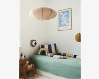 Et hyggeligt børneværelseshjørne med en enkeltseng, der har et lysegrønt, tekstureret sengetæppe. Sengen er pyntet med sjove puder, herunder en stor blå og beige stribet pude, en regnbueformet pude, en blød skyformet pude og en lang, flerfarvet pøllepude. Over sengen hænger en stor, rund, beige papirlampe. Væggen er dekoreret med et indrammet abstrakt kunsttryk i blå og hvid samt en lille dekorativ vifte. En træstol med opbevaringsrum indeholder et tøjdyr og bøger, hvilket tilføjer varme og funktionalitet til rummet.