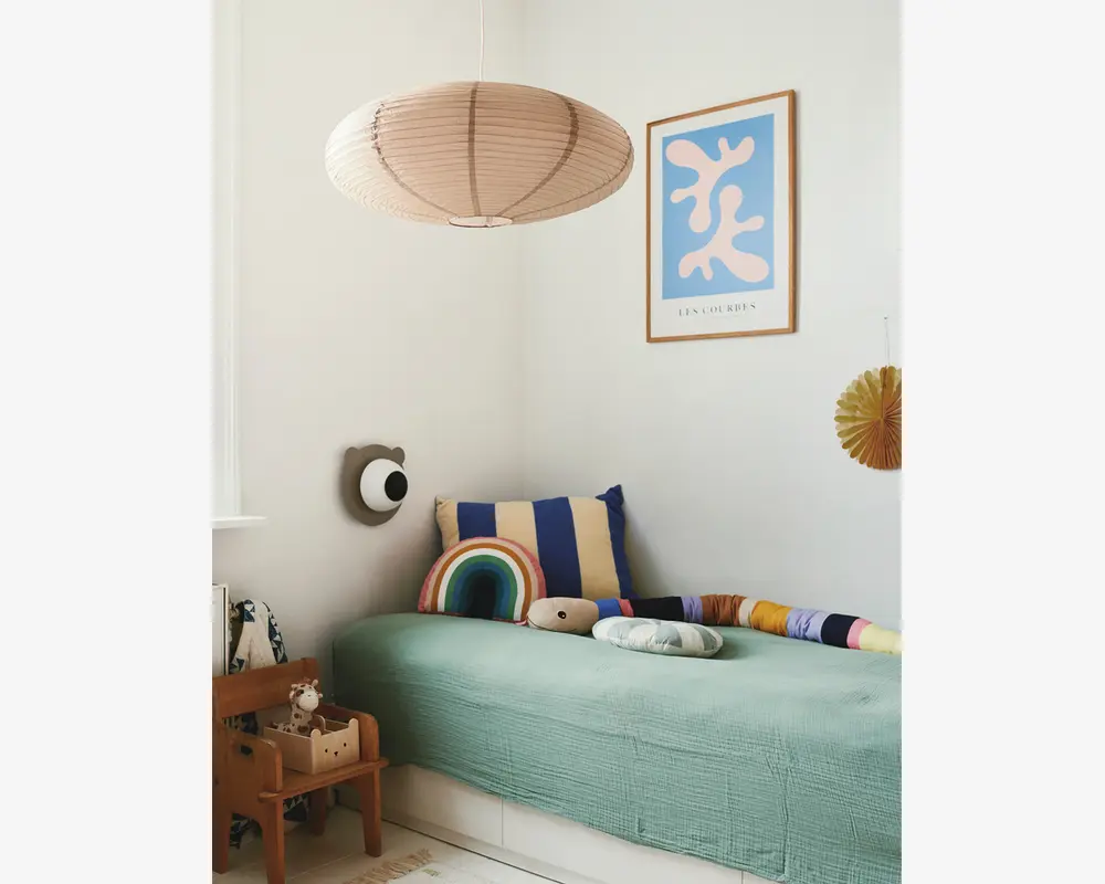 Et hyggeligt børneværelseshjørne med en enkeltseng, der har et lysegrønt, tekstureret sengetæppe. Sengen er pyntet med sjove puder, herunder en stor blå og beige stribet pude, en regnbueformet pude, en blød skyformet pude og en lang, flerfarvet pøllepude. Over sengen hænger en stor, rund, beige papirlampe. Væggen er dekoreret med et indrammet abstrakt kunsttryk i blå og hvid samt en lille dekorativ vifte. En træstol med opbevaringsrum indeholder et tøjdyr og bøger, hvilket tilføjer varme og funktionalitet til rummet.