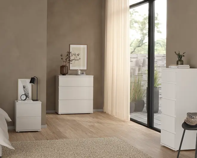 Billedet viser et moderne soveværelse med tre hvide opbevaringsmøbler i forskellige højder. Hvert møbel har et minimalistisk design med glatte skuffefronter uden greb. Den mindste enhed, brugt som sengebord, har to skuffer og er dekoreret med en sort lampe, et indrammet fuglebillede og et lille ur. Den mellemstore enhed har tre skuffer og er pyntet med en brun vase med tørrede grene, et indrammet kunstværk og et lille fad. Den højeste enhed har fem skuffer og er toppet med en lille plante i en brun vase. Alle enheder har en mat finish og rene linjer, der passer til det neutrale rum med trægulv og et stort vindue med lette gardiner.