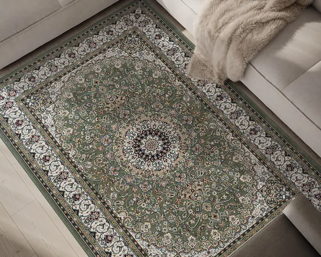 Rektangulært tæppe med et centralt medaljonmønster, der har indviklede blomster- og vinrankemotiver i grønne, cremefarvede, bordeauxrøde og gyldne nuancer. Tæppets kant er dekoreret med udsmykkede motiver i hvid og grøn, hvilket giver et klassisk udtryk. Tæppet har en blød, plysset tekstur og ligger på et lyst trægulv ved siden af en beige sofa med et beige faux pels-tæppe.