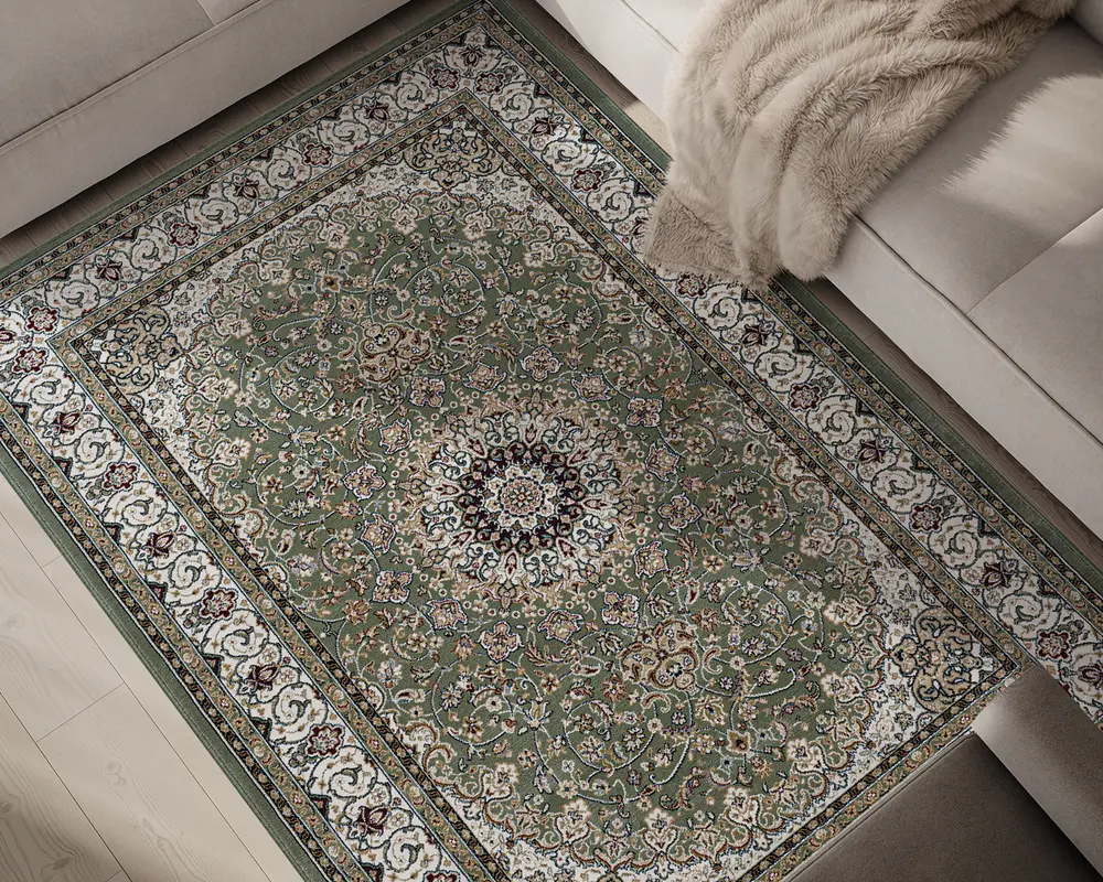 Rektangulært tæppe med et centralt medaljonmønster, der har indviklede blomster- og vinrankemotiver i grønne, cremefarvede, bordeauxrøde og gyldne nuancer. Tæppets kant er dekoreret med udsmykkede motiver i hvid og grøn, hvilket giver et klassisk udtryk. Tæppet har en blød, plysset tekstur og ligger på et lyst trægulv ved siden af en beige sofa med et beige faux pels-tæppe.