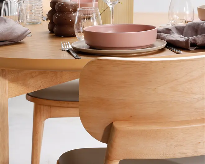 Stol i lyst træ med buet ryglæn og glat finish, kombineret med et matchende rundt spisebord. Stolen har et enkelt, moderne design med let formet sæde for komfort. Borddækningen består af en lyserød keramikskål på en beige tallerken, klart glas, sølvbestik og en sammenfoldet grå serviet, hvilket skaber en moderne og indbydende spiseatmosfære.