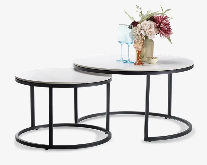 Sæt med to runde indskudsborde med hvide bordplader og sorte metalstel. Det større bord er pyntet med en vase med kunstige blomster, to blåtonede glas og en lille dekorativ skål. Bordene har et moderne, minimalistisk design med åbne cirkulære baser, så det mindre bord kan skubbes delvist ind under det større for at spare plads.