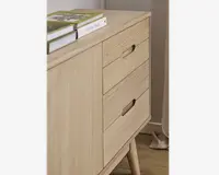 Skab i lyst træ med tre skuffer, der har integrerede greb, samt en enkelt låge på venstre side. Skabet står på skråtstillede ben og har en glat, naturlig finish. Der ligger flere bøger ovenpå, og designet er minimalistisk og moderne.