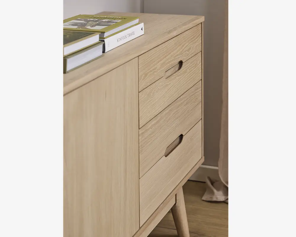 Skab i lyst træ med tre skuffer, der har integrerede greb, samt en enkelt låge på venstre side. Skabet står på skråtstillede ben og har en glat, naturlig finish. Der ligger flere bøger ovenpå, og designet er minimalistisk og moderne.
