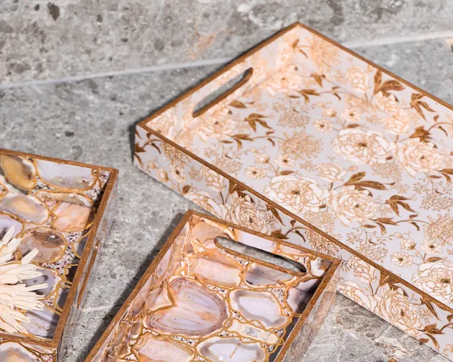 Sæt af dekorative rektangulære bakker med udskårne håndtag. Én bakke har et guld- og hvidt blomsterdesign på indersiden og siderne, mens de andre har et marmoreret agatstensmønster i nuancer af beige, brun og lavendel med gulddetaljer. Bakkerne har en blank overflade og forhøjede kanter for sikker transport.