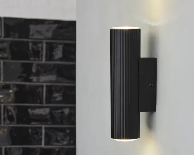 Et moderne vægmonteret lysarmatur med en lodret cylindrisk form, udført i mat sort. Cylinderen har diskrete lodrette riller, der giver ekstra tekstur. Lyset udsendes både fra toppen og bunden, hvilket skaber en op- og nedadgående lyseffekt på væggen. Armaturet er fastgjort til væggen med en rektangulær sort monteringsplade.